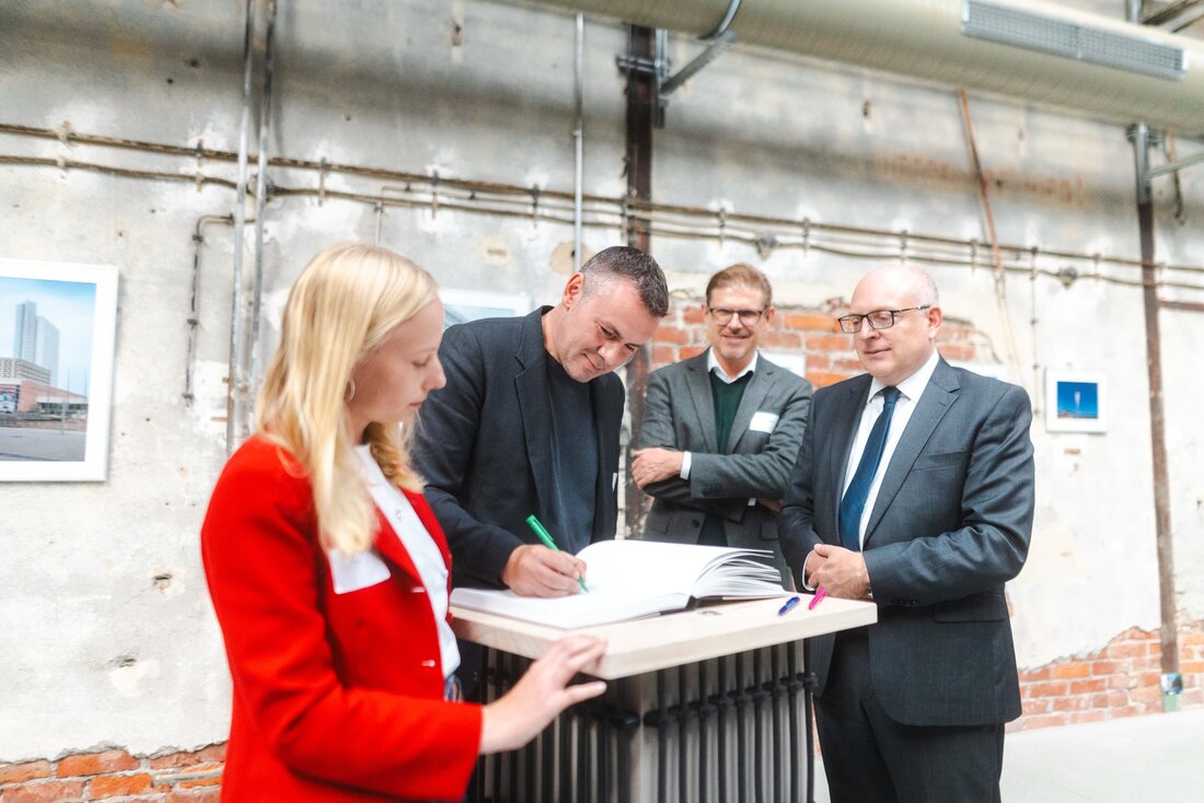 Zu sehen sind vier Personen, die an einem Tisch stehen. Eine Person unterschreibt etwas in einem Buch, zwei Personen stehen direkt an dem Tisch, die vierte Person steht etwas versetzt im Hintergrund und hat die Arme verschränkt. Alle Personen lächeln. Sie stehen in einer umgebauten Industriehalle. 