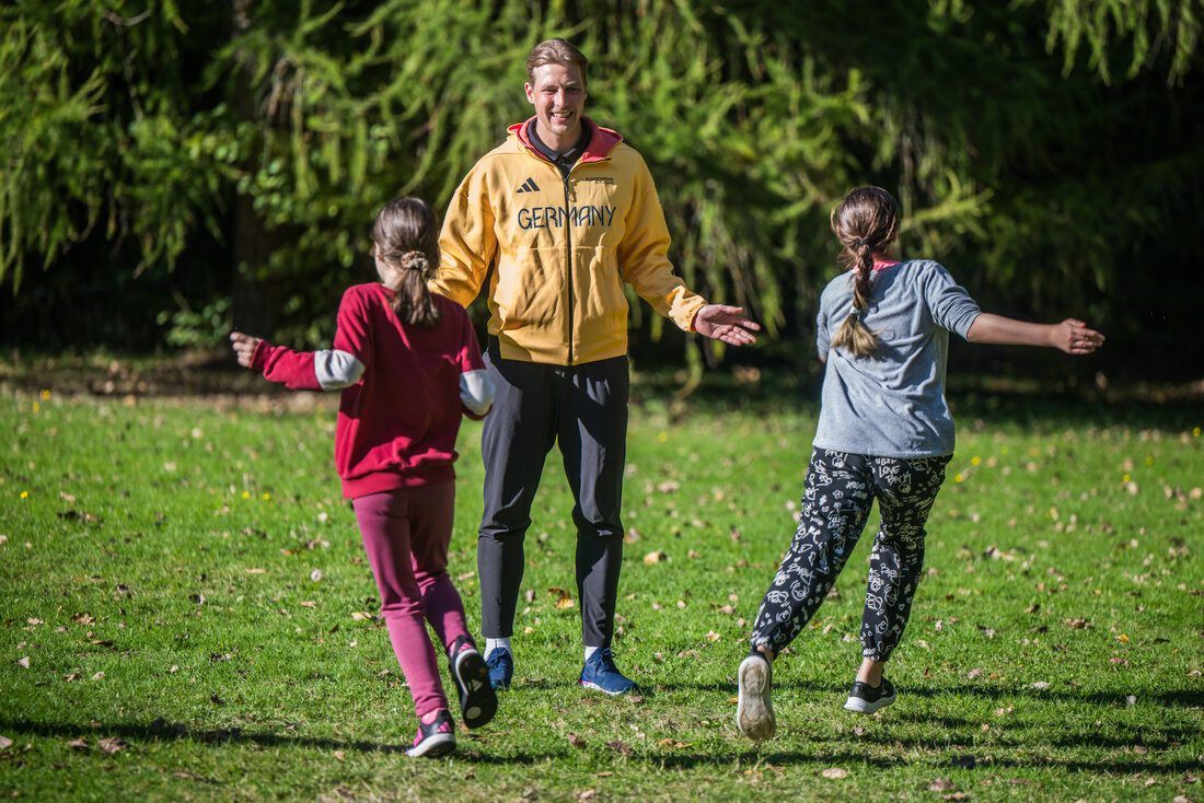 Der Weitspringer Max Heß in einer gelben Trainings-Jacke lächelt und heißt zwei Kinder mit offenen Armen auf einer grünen Wiese willkommen.