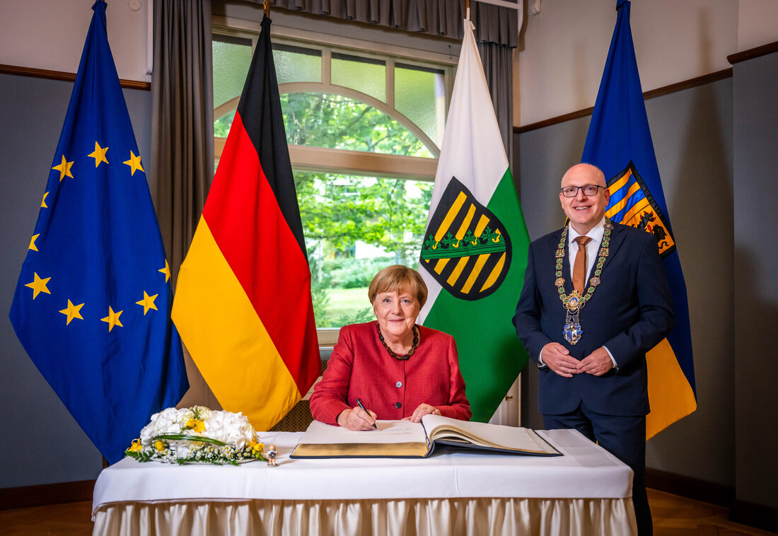 In der Mitte des Bildes sitzt Angela Merkel hinter einem Tisch. Sie trägt einen roten Blazer mit schwarzen Knöpfen sowie eine Halskette mit dunklen Bernsteinen. In der rechten Hand hält sie einen Stift, mit dem sie im Goldenen Buch der Stadt Chemnitz unterschreibt. Auf der linken Seite des Tisches steht ein Blumengesteck in weiß und gelb sowie ein kleiner Engel mit einem Zahnrad in den Händen. Rechts neben ihr steht Oberbürgermeister Sven Schulze. Er trägt einen dunkelblauen Anzug, ein weißes Hemd, eine kupferfarbene Krawatte sowie seine Amtskette. Im Hintergrund sind vor einem Fenster die Fahnen von Europa, Deutschland, Sachsen und der Stadt Chemnitz aufgestellt.