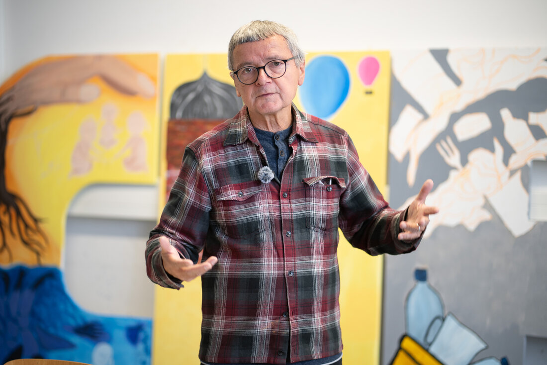 Gerald Richter, making an explanatory gesture in front of colourful, large-scale paintings in a gallery or studio.