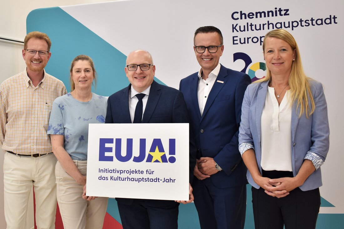You can see three men and two women standing next to each other in front of a wall. They are all smiling at the camera. One man is holding a sign that reads EUJA! 