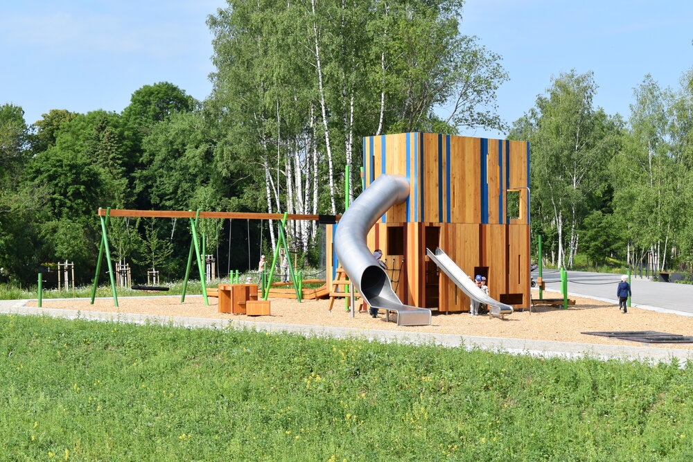 Im Vordergrund eine Wiese, dahinter ein großer Spielplatz mit Kletterturm und Rutsche, im Hintergrund Bäume.