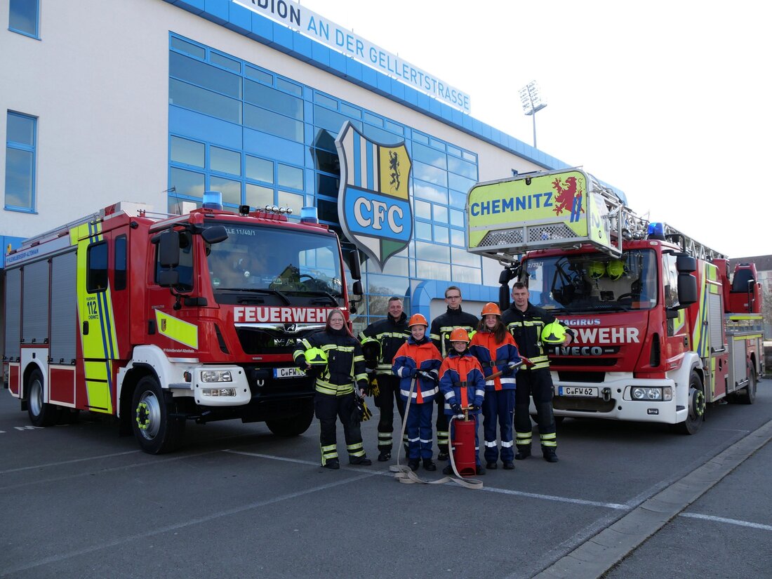 Im Bild sind zwei Feuerwehr-Einsatzwagen zu sehen, eines mit und eines ohne Drehleiter. Dazwischen vorn in der Mitte seht eine Gruppe Feuerwehrleute, eine Frau und drei Männer. Vor ihnen drei Kinder der Feuerwehrjugend. Alle haben Feuerwehrkleidung an. Sie schauen frontal in die Kamera. Im Hintergrund sieht man das Stadion an der Gellertstraße. In der Mitte prangt das CFC-Wappen.