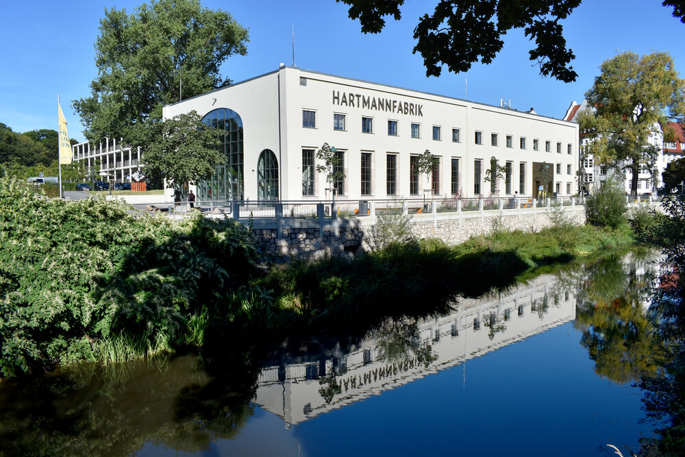Die Hartmannfabrik spiegelt sich im Fluss Chemnitz, umrahmt von grünen Bäumen.