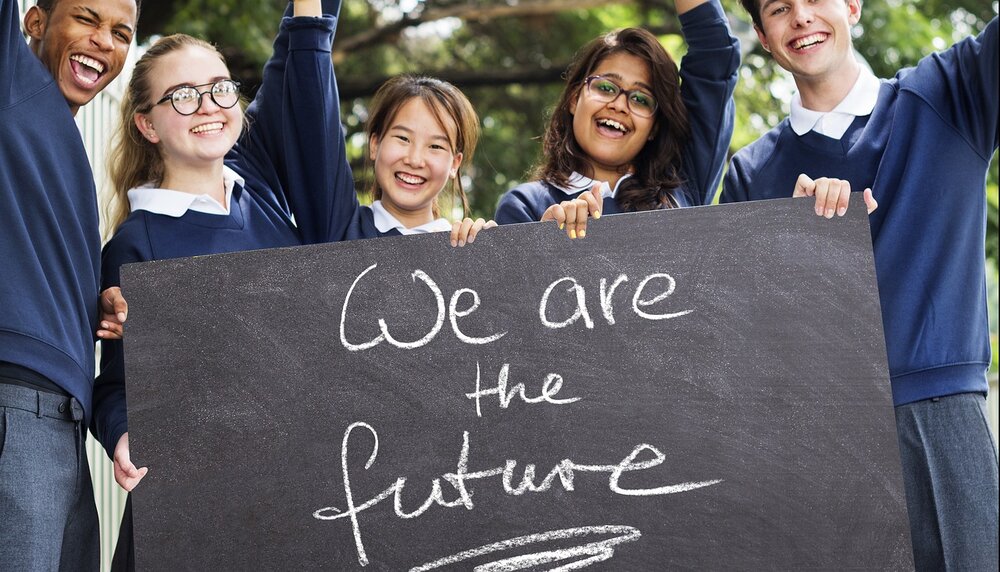 Schülerinnen und Schüler halten eine Tafel vor sich mit der Aufschrift "We are the future", sie lächeln und strecken die Arme hoch