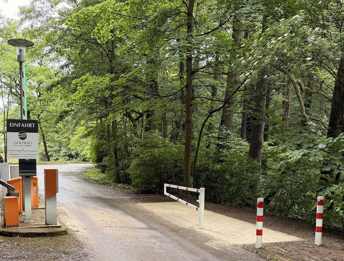 Auf dem Bild ist eine Zufahrt zu sehen, deren Schranke offen steht. Neben der Schranke ist eine sandfarbene befestigte Fläche, die es Fußgängern, Rad- und Rollstuhlfahrern auch bei geschlossener Schranke ermöglicht, die Zufahrt zu passieren, davor sind rot-weiße Poller, um eine Durchfahrt von PKW zu verhindern. Im Hintergrund sind dichte Bäume zu sehen.