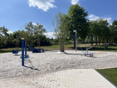 Im Fokus ist der Spielplatz. Verschiedene Spielgeräte stehen auf einem Sandigen Untergrund. Im Hintegrund sieht man mehrere ältere grüne Bäume.