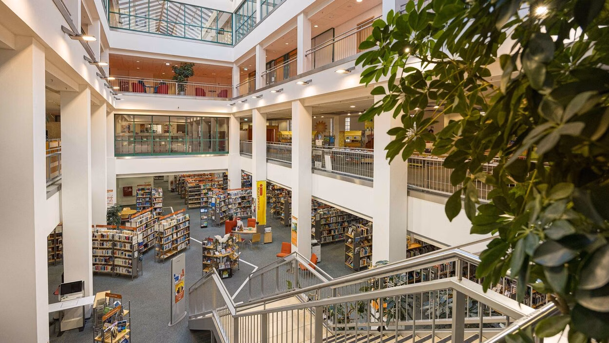Innenansicht der Zentralbibliothek der Stadtbibliothek: Ein lichtdurchfluteter, mehrstöckiger Raum mit weißen Säulen, offenen Galerien und zahlreichen Bücherregalen im Erdgeschoss, überdacht von einem großen Glasdach.