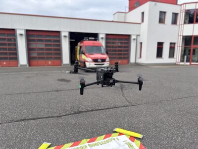 Das Bild zeigt eine Drohne, die in der Luft etwa einen Meter &uuml;ber dem Boden schwebt. Der Boden besteht aus Asphalt, auf dem Asphalt liegt ein textiler Landeplatz. Im Hintergrund ist eine Feuerwache und ein Feuerwehrauto zu sehen.