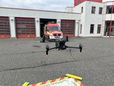 Das Bild zeigt eine Drohne, die in der Luft etwa einen Meter über dem Boden schwebt. Der Boden besteht aus Asphalt, auf dem Asphalt liegt ein textiler Landeplatz. Im Hintergrund ist eine Feuerwache und ein Feuerwehrauto zu sehen.