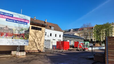 Auf dem Bild sind Gebäude zu sehen, davor befinden sich Baucontainer und unbefestigter Erdboden. Im Vordergrund steht ein bauschils, auf dem steht: Wir schaffen - die Kulturhauptstadt Europas 2025 - Räume für MaherInnen. Im Hintergrund sind Wohnhäuser des Sonnenbergs zu sehen und ein strahlend blauer Himmel.