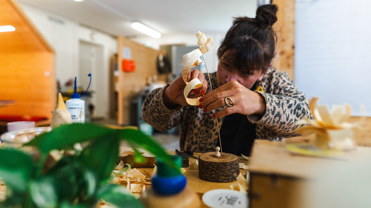 In einer rustikalen Werkstatt arbeitet eine Person an einem Tisch mit Holz. Auf der Werkbank liegen zahlreiche feine Holzspäne und verschiedene Werkzeuge. Im Hintergrund sind weitere Holzarbeiten und Materialien zu sehen. Die warme Beleuchtung und die hölzerne Einrichtung vermitteln die Atmosphäre einer traditionellen Handwerkswerkstatt.