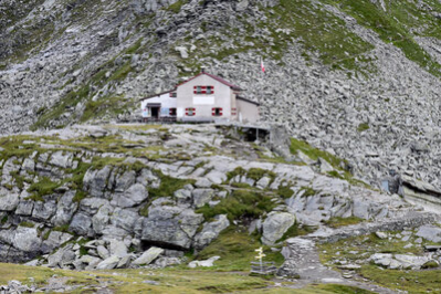 Das Bild zeigt von Weiten die Chemnitzer Hütte. Die Hütte ist umgeben von massiven Felsen. Zwischen den Felsen ist etwas Wiese zu erkennen. Die Chemnitzer Hütte ist ein graufarbenes Gebäude mit rotweisen Fensterklappläden. Vor dem Haus ist ein kleiner Sitzbereich zu sehen. Rechts neben dem Haus befindet sich ein Fahnenmast mit einer Flagge. Diese ist jedoch nicht zu erkennen, da kein Wind weht.