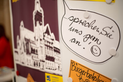 A cut-out speech bubble hangs on a pinboard next to the poster for the residents' meeting. The speech bubble contains the text "Feel free to contact us!"