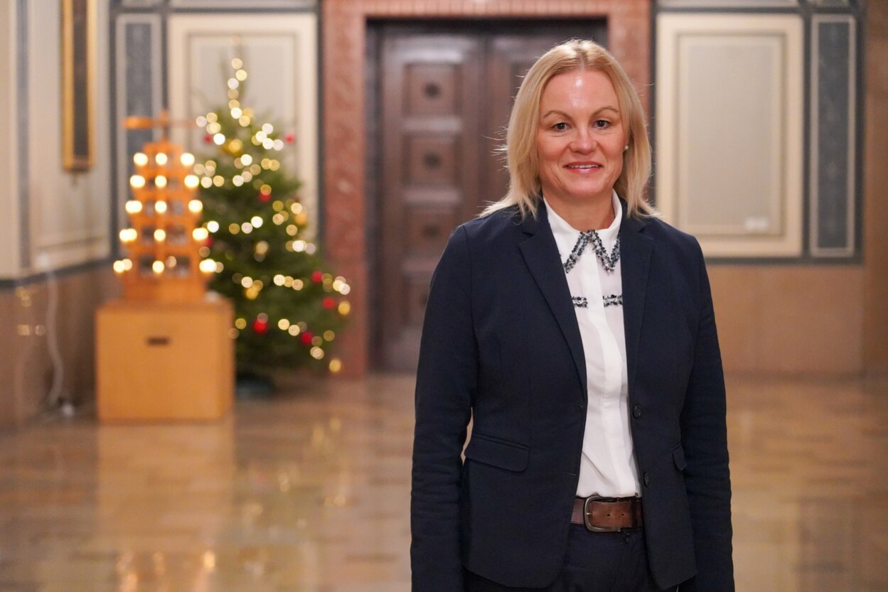 Im Bild ist eine Frau vom Kopf bis zur Hüfte zu sehen. Sie trägt halblange blonde Haare, ein dunkles Sacko und eine weiße Bluse darunter. Sie steht in der Wandelhalle im Rathaus, im Hintergrund steht ein Weihnachtsbaum und eine Weihnachts-Pyramide, beide beleuchtet.