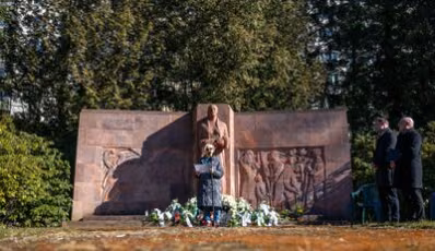 Das Bild zeigt das Mahnmal auf dem Städtischen Friedhof. Davor steht die Zeitzeugin Eva Otto an einem Mikrophon. In der Hand hält sie ihre Rede. Sie trägt einen langen dunklen Mantel und eine Sonnenbrille, da die Sonne scheint.