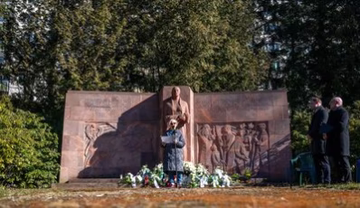 Das Bild zeigt das Mahnmal auf dem Städtischen Friedhof. Davor steht die Zeitzeugin Eva Otto an einem Mikrophon. In der Hand hält sie ihre Rede. Sie trägt einen langen dunklen Mantel und eine Sonnenbrille, da die Sonne scheint.