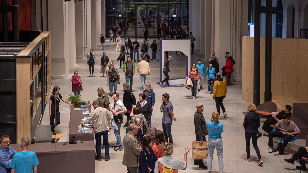 Innenansicht der Hartmannfabrik aus erh&ouml;hter Perspektive von einer oberen Etage, mit Blick in die gro&szlig;e Industriehalle, in der sich zahlreiche Besucher zwischen Ausstellungsbereichen und offenen Fl&auml;chen bewegen.