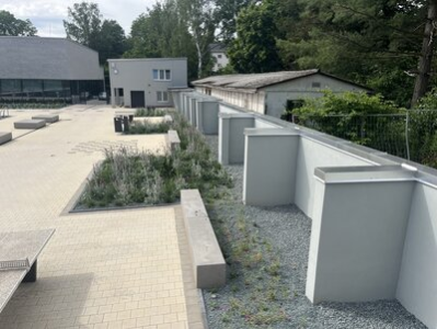 Das Bild zeigt den Spielbereich neben dem 50 Meter Schwimmbecken. Dort befinden sich 2 Tischtennisplatten und eine Fläche zum Schach spielen. An der Mauer, welche das Freibad eingrenzt, befindet sich ein striefen Kieß mit verschiedenen grünen Pflanzen. Im Hintergrund sieht man einen kleinen Teil des Hallenbades.