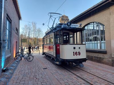 Im Bild ist ein historischer Stra&szlig;enbahnwagong mit beiger Lackierung zu sehen, er steht auf einer Schiene, die von Pflastersteinen umgeben ist. Links und rechts davon befinden sich Geb&auml;ude des garagen-Campus. Im Hintergrund sind Menschen zu sehen, die zur Er&ouml;ffnung das Gel&auml;nde besuchen, und ein strahlend blauer Himmel..