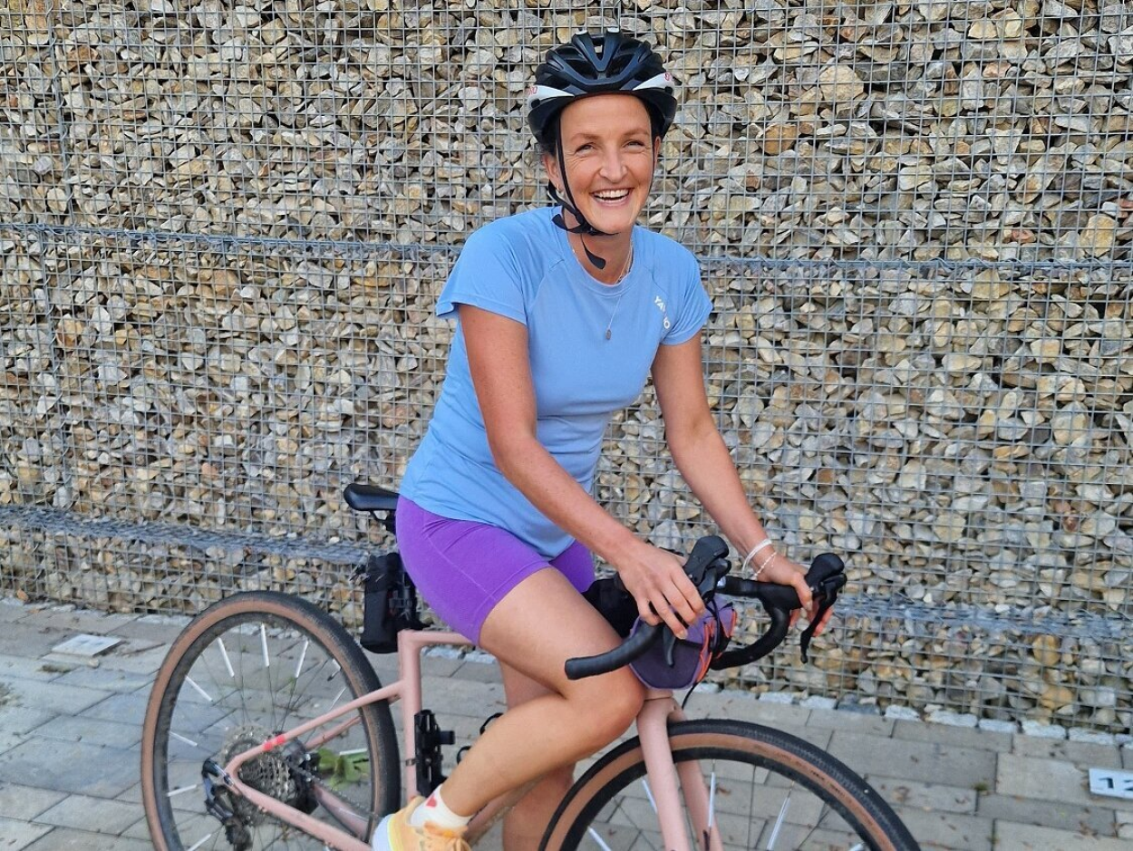 Im bild ist eine Radfahrerin zu sehen. Sie steht mit einem Bein auf dem Boden, sitzt aber im Sattel und hat die Hände am Lenkrad. Sie lächelt in die Kamera, trägt einen Helm, ein blaues Shirt und lilafarbene Radlerhosen. Sie steht auf einem Grund aus verlegten Granitplatten, im Hintergrund ist eine Gabionen-Wand (Steine im Gitter)