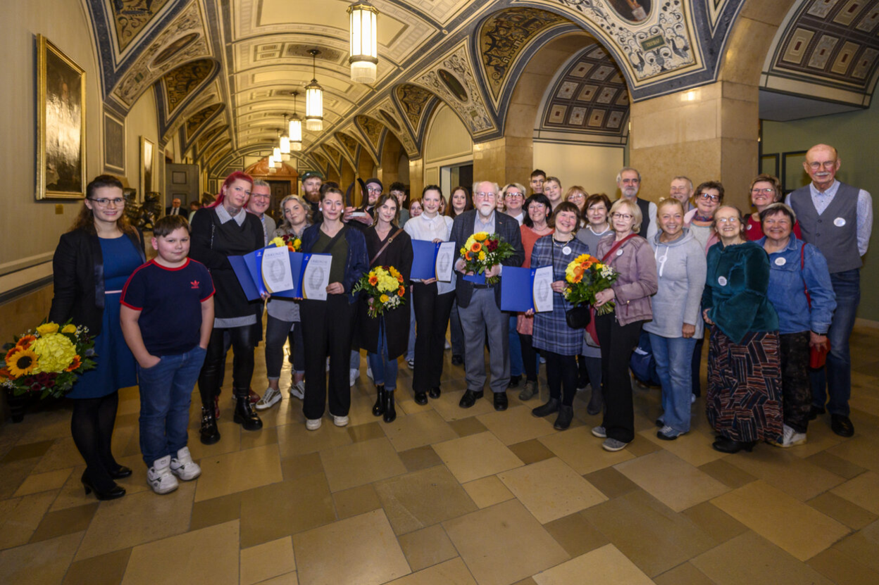 In der Wandelhalle des Rathauses stehen die vielen Preisträgerinnen und Preisträger. Sie halten ihre Urkunden und Blumen in den Händen.