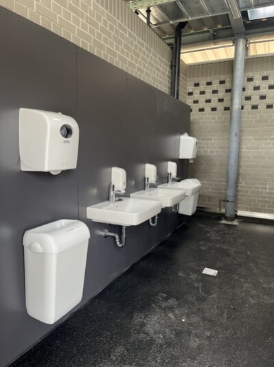 Das Bild zeigt 3 Waschbecken. Links und rechts neben den 3 Waschbecken hängt ein Mülleimer und ein Papierhalter.