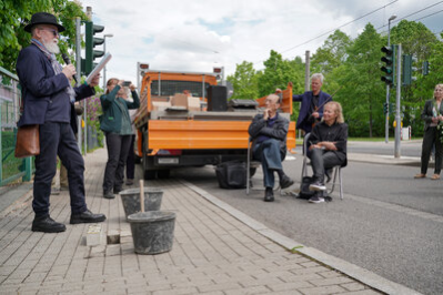 Dr. Nitsche hält eine Rede und blickt zu den Besucherinnen und Besuchern; Angehörige sitzen auf Stühlen und schauen ihm zu. Im Hintergrund ist ein Baustellenfahrzeug zu sehen.