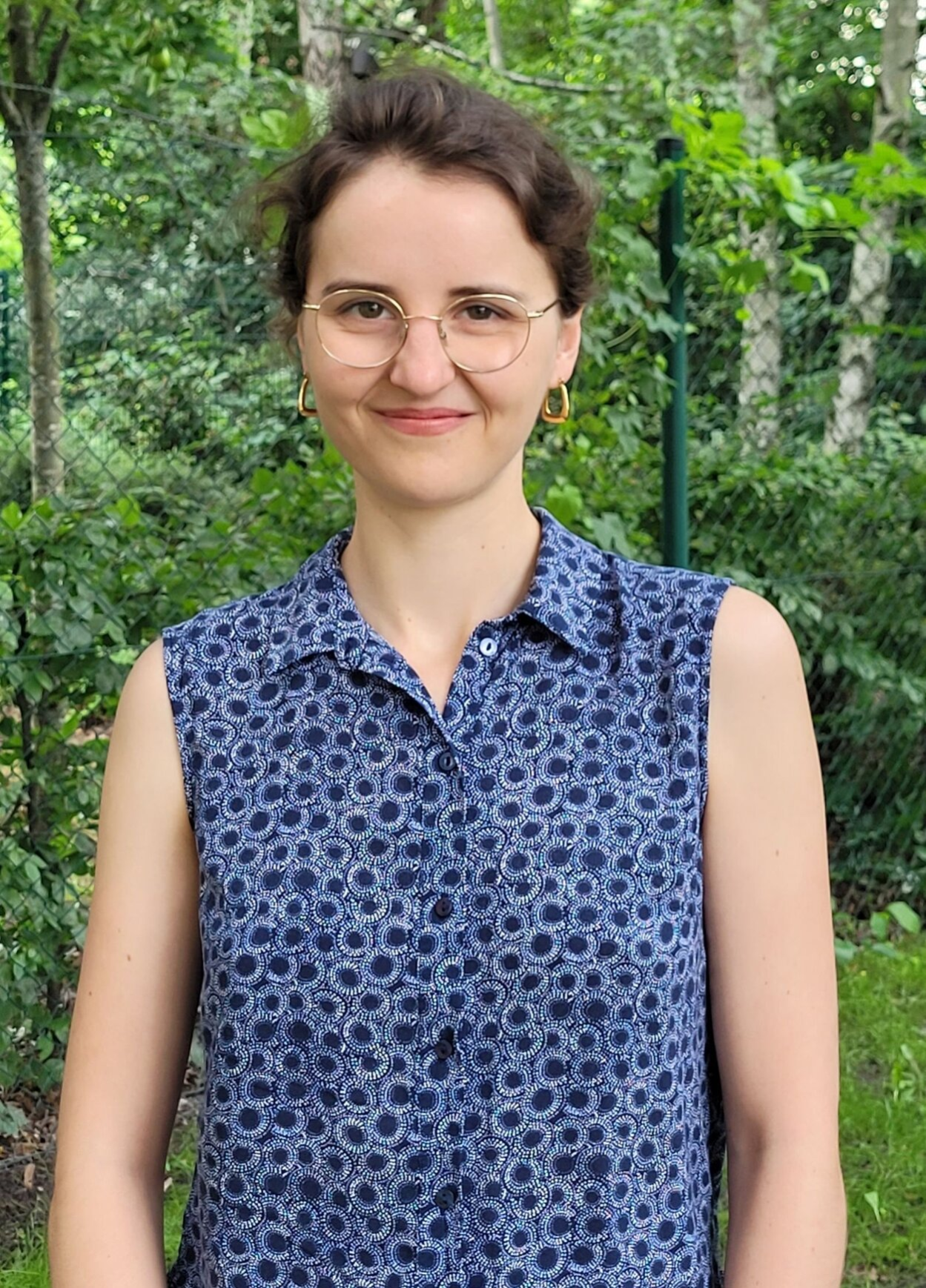 Im Bild ist eine junge Frau zu sehen, sie hat braune Haare zu einem Zopf zusammengebunden und trägt eine Brille sowie eine ärmellose Bluse mit einem dunkelblauen Muster. Sie steht im Freien, hinter ihr sind grüne Bäume und Sträucher zu sehen.