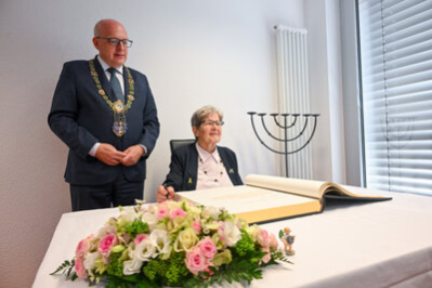 Renate Aris trägt sich anlässlich ihres 90. Geburtstags in das Goldene Buch der Stadt Chemnitz ein. Neben ihr steht Oberbürgermeister Sven Schulze mit Amtskette. Auf dem Tisch vor ihr liegt das große aufgeschlagene Buch, geschmückt mit einem Blumenstrauß. Im Hintergrund ist ein siebenarmiger Leuchter zu sehen.
