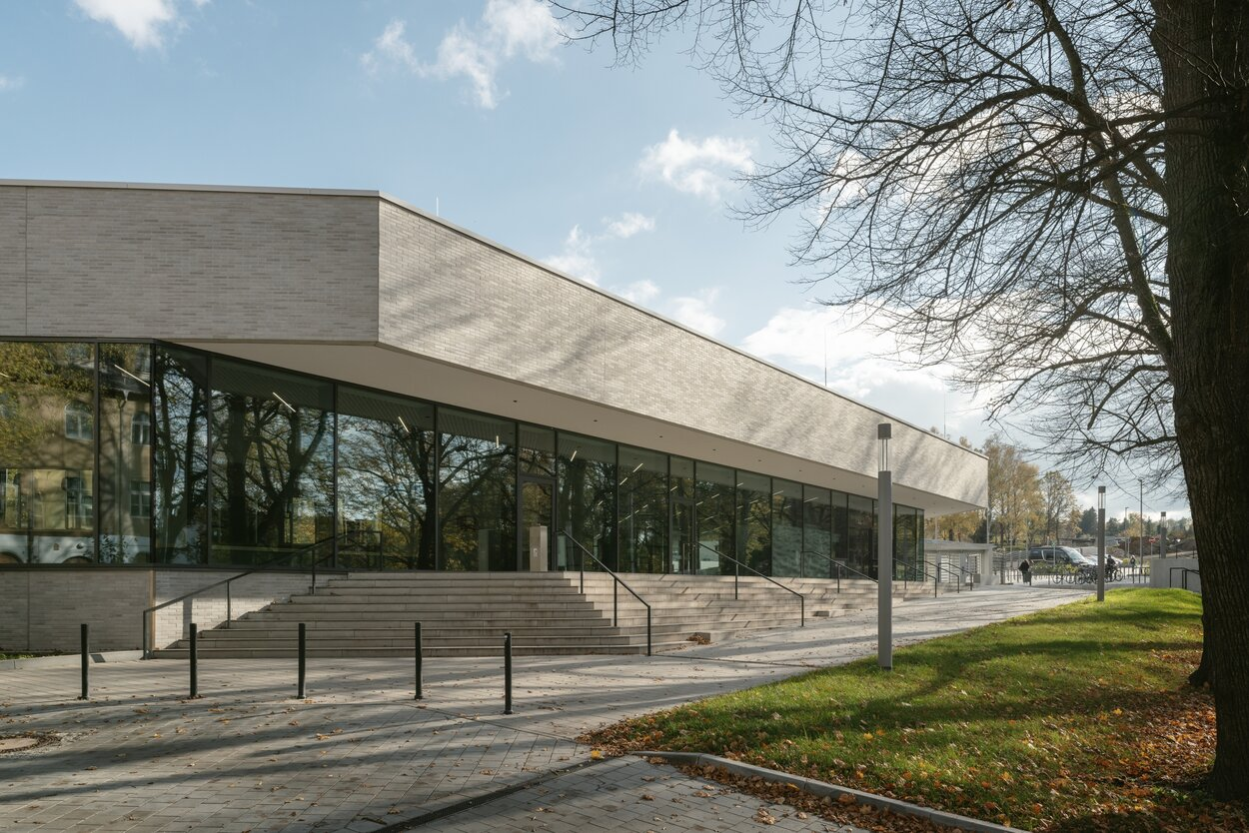Außenansicht der Schwimmhalle Bernsdorfer Bad: Die Front ist voll verglast. Davor befinden sich Treppen mit Geländer, auf der rechten Seite führt ein barrierefreier Weg zum Eingang.