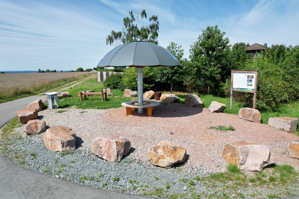 Runder Wetterpilz mit Sitzbank, umgeben von rund 20 großen Findlingen, im Hintergrund ein weites Feld.