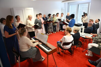 Ein Chor singt in einem hellen Raum, begleitet von einer Pianistin am Keyboard. Vor dem Chor sitzt das Publikum auf Stühlen, darunter Renate Aris und Oberbürgermeister Sven Schulze.