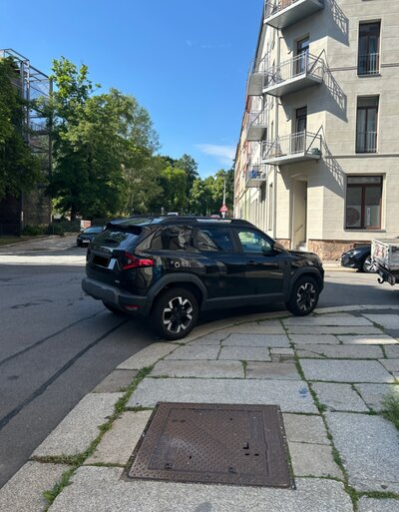 Ein schwarzes Auto steht in der Kurve einer Kreuzung halb auf der Fahrbahn und halb auf dem Gehweg.