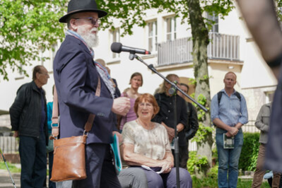 Dr. Nitsche hält eine Rede, während Tana Sachs aufmerksam zuhört; im Hintergrund sind Besucherinnen und Besucher der Stolpersteinverlegung zu sehen.