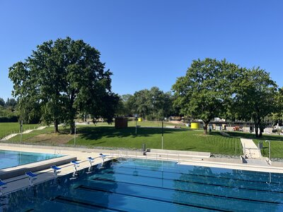Das Bild zeigt im Vordergrund das Schwimmbecken. Im Hintergrund erkennt man die Liegewiese mit großen grünen Bäumen.