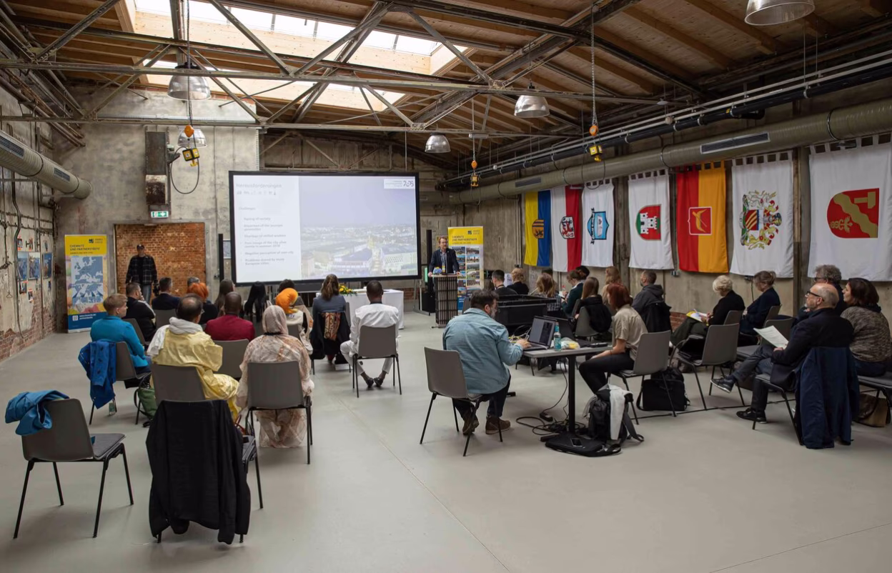 In einer gro&szlig;en, ehemaligen Fabrikhalle, sitzt eine Gruppe an Menschen und lauschen einem Vortrag, der stattfindet. Es h&auml;ngen mehrere Flaggen an einer Wand auf der rechten Seite.
