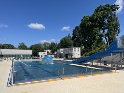 Zu sehen ist das Mehrzweckbecken mit der Wellenrutsche. Im Hintergrund befinden sich die Gebäude des Hallenbades.