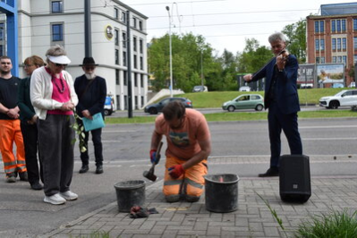 Ein Bauarbeiter verlegt die Stolpersteine; links von ihm stehen Sonia Ascher und Besucherinnen und Besucher, rechts ist ein Musiker zu sehen.