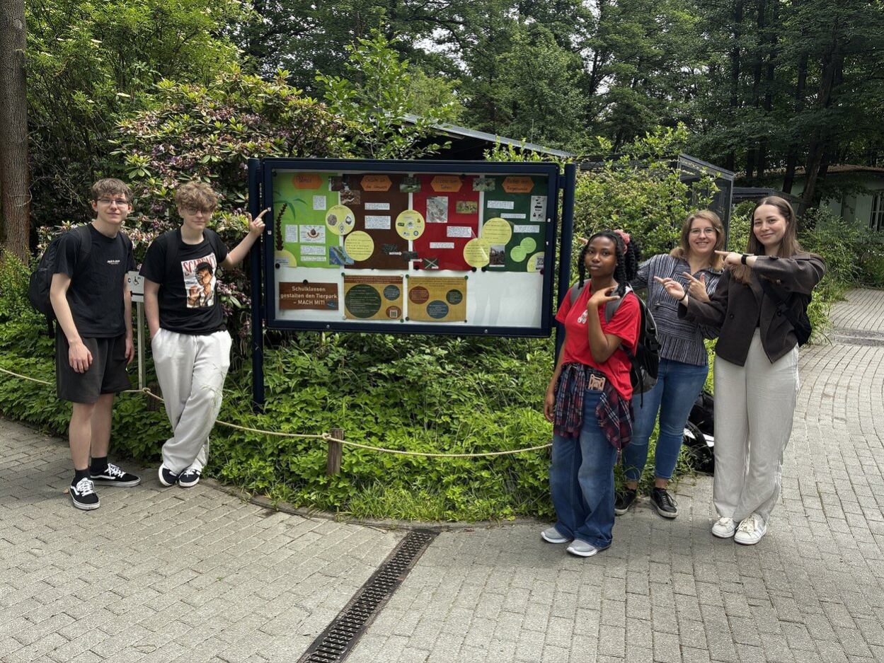 Eine Gruppe an Jugendlichen stehen rechts und links eeines Schaukastens und lächeln in die Kamera.