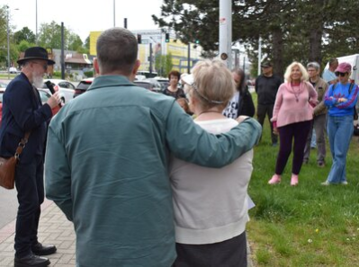 Sonia Ascher wird von ihrem Mann im Arm gehalten; im Hintergrund sind Besucherinnen und Besucher zu sehen, während Dr. Nitsche seine Rede hält.