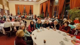 A group of senior citizens are sitting at several large tables with drinks in front of them. They are chatting with interest.