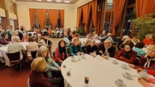 Eine Gruppe Seniorinnen und Senioren sitzen an mehreren großen Tischen mit Getränken vor sich. Dabei unterhalten sie sich interessiert.