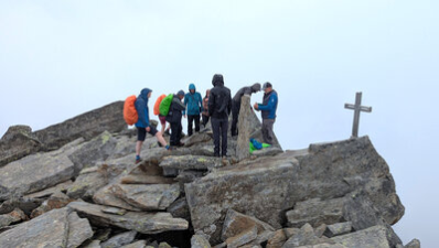 Das Bild zeigt mehrere Personen auf Felsen am Gipfelkreuz. Die Felsen sind grau und sehen massiv aus. Alle Peronen tragen Wanderkleidung. Außerdem tragen alle Personen eine Kaputze auf dem Kopf. Im Hintergrund ist nur grauer Nebel zu erkennen.