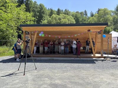 Choir sings in an open hiker's hut at the edge of the forest, in the foreground is a tripod with a video camera for recording.