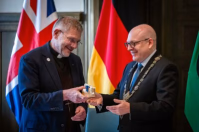 The Very Reverend Dean John Witcombe, Vorsteher der Coventry Gemeinde und Oberbürgermeister Sven Schulze stehen vor der britischen und deutschen Flagge. Der Oberbürgermeister trägt die Amtskette und übergibt The Very Reverend Dean John Witcombe ein braunes Kästchen.