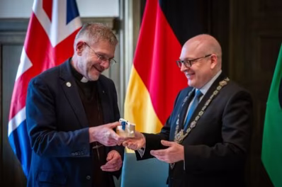 The Very Reverend Dean John Witcombe, Vorsteher der Coventry Gemeinde und Oberbürgermeister Sven Schulze stehen vor der britischen und deutschen Flagge. Der Oberbürgermeister trägt die Amtskette und übergibt The Very Reverend Dean John Witcombe ein braunes Kästchen.