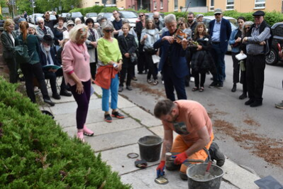 Ein Bauarbeiter verlegt den Stolperstein; im Hintergrund beobachten Besucherinnen und Besucher die Verlegung, während ein Musiker spielt