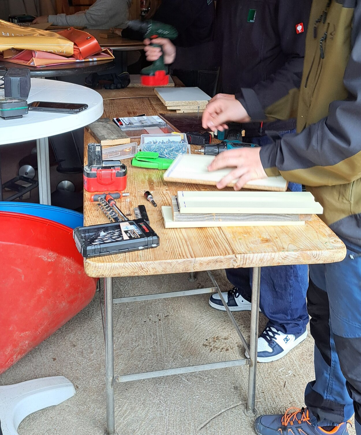 Auf einem Biertisch liegen Werkzeuge und Holzbretter. Jugendliche halten einen Akkuschrauber und ein Holzbrett in der Hand.