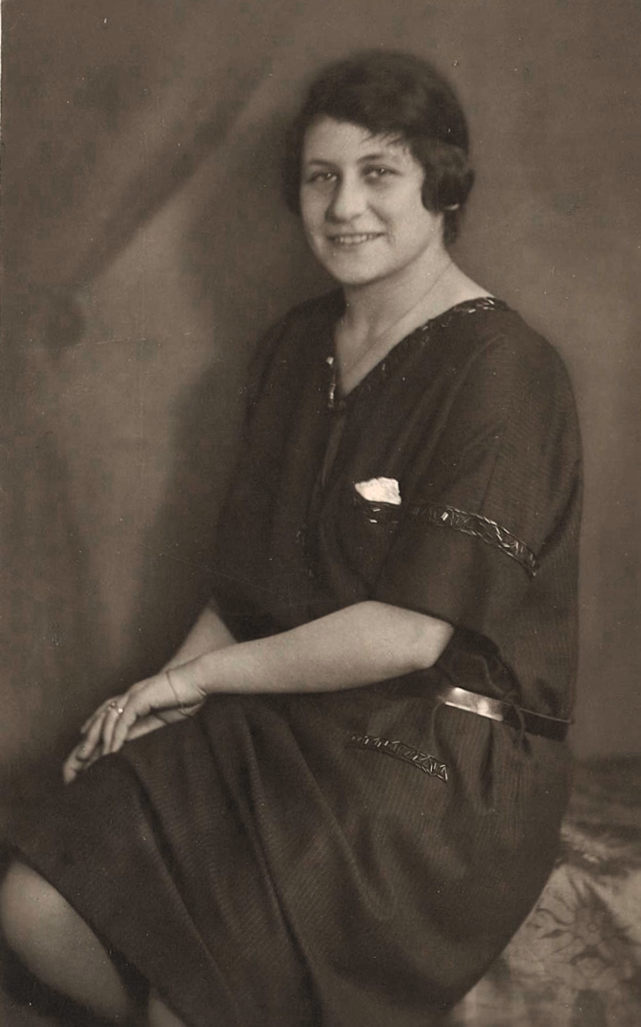 Schwarz-Weiß-Foto von Liselotte Firnbacher. Im Sitzen hat sie die Hände auf ihrem rechten Bein abgelegt und trägt ein weites, langes Kleid. Sie lächelt in die Kamera und trägt ihre Haare kurz.